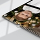 Close-up of an A4 wall calendar with a sleek black design, showing a photo of a young child in front of a Christmas tree. The calendar has spiral binding with a metal hanging hook and part of a monthly grid visible.