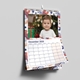 Wall calendar open to December 2026, featuring a young child in front of a decorated Christmas tree with presents. The page has a festive abstract border in red, blue, and beige, and includes a clear monthly grid with bold dates and weekdays. The calendar hangs neatly on a white wall using a metal hook