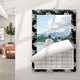 Wall calendar with a floral border featuring a scenic photo of two people in winter clothing gazing at snow-capped mountains across a lake. The calendar is mounted on a white wall in a bright, modern living space with natural light streaming in. The bottom page is partially turned, revealing the dated grid below.