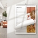 A spiral-bound wall calendar open to July 2025, displayed in a modern living room. The page shows space for notes alongside a photo of a young child sitting on straw next to a wooden door. The calendar hangs on a light-coloured wall.