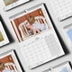 An A4 wall calendar for July 2025 with a marble design, featuring a photo of a young child gently kissing a baby on a rustic bench. The calendar has a monthly grid, spiral binding, and a hanging hook. Multiple calendar designs are displayed underneath.