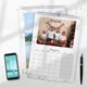 A4 wall calendar with a marble design, open to December 2025. The page features a family Christmas photo with festive decorations. A second calendar showing a lake and mountain scene is partially visible underneath. A smartphone and pen are placed beside the calendars.