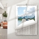 A 2026 wall calendar with a white marble border hangs on a light-coloured wall in a modern living space. The page is partially turned, revealing a scenic photo of two people standing by a lake, looking at snow-covered mountains under a cloudy sky. The calendar is spiral-bound with a central hanging hook, and the visible grid shows the end of a monthly layout. A cosy lounge area with natural light appears in the background.