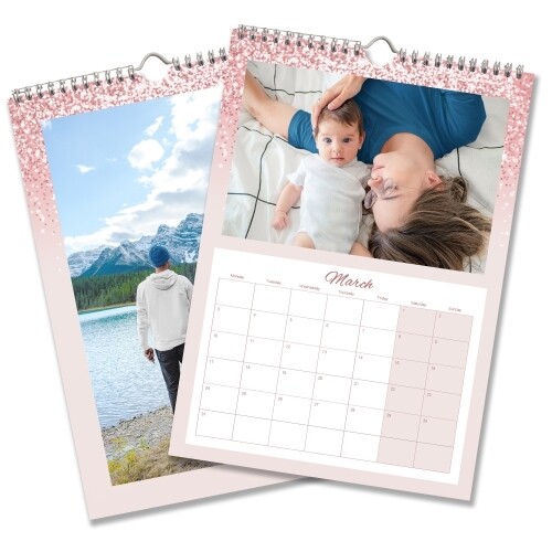 Two A3 wall calendars with pink glitter borders. One features a landscape photo of a person by a lake and mountains, and the other shows March 2025 with a photo of a parent and baby lying on a bed. Both calendars have monthly grids, spiral bindings, and hanging hooks.