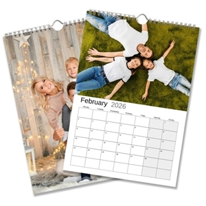 February 2026 wall calendar featuring a photo of a family lying on green grass, smiling and dressed in matching white tops and jeans. Behind it is another page with a festive family portrait against a rustic wooden backdrop with warm fairy lights.