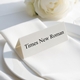 Personalised wedding place card with your name in elegant black script on folded white cardstock. Positioned on a neatly folded white napkin atop a white plate, with a soft-focus white rose in the background.