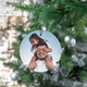 Personalised ceramic bauble hanging on a Christmas tree, featuring a playful image of a young girl sitting on an adult man's shoulders, covering his eyes. The background has festive greenery and silver ornaments.