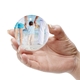 Hand holding a personalised ceramic bauble featuring an image of a child at the beach, holding hands with two adults. The child faces the ocean while one adult holds a teddy bear, creating a heartwarming family scene.