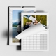 Two open wall calendars are shown laid out on a white surface. The front calendar displays a photo of a happy family lying on grass, with the page partly turned to reveal a blank monthly grid. The calendar behind features a person in a white hoodie standing by a lake with mountains and trees in the background. Both calendars have a white design with wire binding and a central hanging hook.