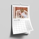 Wall calendar for February 2026 hanging on a grey wall. The top half features a heart-warming photo of a young child lovingly cuddling a baby on a distressed white bench. The calendar has a clean white design with spiral binding and a hanging hook, with the bottom page curling slightly forward.