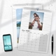 Two A4 photo calendars for 2025, one opened to May. The May calendar features your own image. A smartphone and pen are placed next to the calendars on a white desk, with space for notes in the month grids.