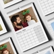 Flat lay of multiple wall calendars featuring personal photos, with the main focus on the December 2026 page showing a bearded man holding a smiling baby wearing green reindeer antlers. The calendar has a clean grid layout with a spiral binding and hanging hook, surrounded by other pages showing different months and family moments.
