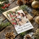 A personalised Christmas card featuring a smiling family of three in festive jumpers, with the man holding a baby on his shoulders and the woman beside them. The card is placed on a gift surrounded by pine branches, pinecones, and gold baubles. It reads, 