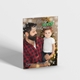 A Christmas card featuring a man with a beard in a red flannel shirt holding a young child wearing reindeer antlers. The background shows a lit Christmas tree, and the card reads, 