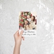 A hand holding a personalised Christmas card featuring a family of three. The card shows a man carrying a baby on his shoulders with a smiling woman next to them, all in front of a decorated Christmas tree. The message reads, 