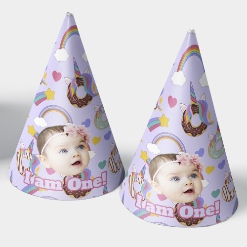 Two customised party hats with pastel purple designs featuring rainbows, stars, donuts, and unicorn details. Each hat includes a personalised photo of a baby wearing a pink flower headband and the text I am One, ideal for a first birthday celebration.