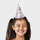 Smiling girl wearing a customised party hat with a pastel purple design featuring rainbows, stars, donuts, and unicorns. The hat includes a personalised baby photo and the text I am One, perfect for a first birthday celebration.