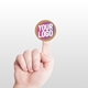 large kraft circle label sticker with a custom pink and white logo, placed on the tip of an extended index finger. The minimalist white background accentuates the personalised design and eco-friendly material