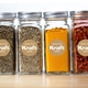 Four glass jars with silver lids filled with herbs, spices and seeds, each labelled with a round 25mm personalised Kraft label featuring white print. The custom brown stickers display the text White Print Kraft Labels and add a rustic touch to kitchen storage or pantry organisation.