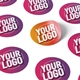 Personalised 25mm round labels with a custom logo design. The stickers are printed on a sheet and come in various shades of pink, purple, and orange. One label is partially peeled to show the adhesive backing. Ideal for branding, packaging, or crafts.