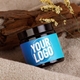 A dark amber glass jar with a black screw-on lid, featuring a blue custom label with a placeholder for a logo. The jar is placed on a soft beige towel with dried flowers and a wooden accent in the background, creating a natural and spa-like setting. Ideal for personalised branding of skincare products, creams, balms, or handmade cosmetics. The amber glass helps protect contents from light exposure.