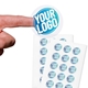 Transparent custom circle label sticker with a blue and white logo, shown on a fingertip, alongside two sheets filled with multiple identical stickers. Ideal for personalised branding or product labelling.