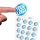 Transparent custom circle label sticker with a blue and white logo, shown on a fingertip, alongside two sheets filled with multiple identical stickers. Ideal for personalised branding or product labelling.