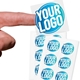 Transparent custom square label sticker with a blue and white logo, shown on a fingertip, alongside two sheets filled with multiple identical stickers. Ideal for personalised branding or product labelling.