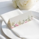 wedding place card with an elegant design on folded white cardstock, ready to write your names on. Positioned on a neatly folded white napkin atop a white plate, with a soft-focus white rose in the background.