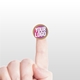 Small kraft circle label sticker with a custom pink and white logo, placed on the tip of an extended index finger. The minimalist white background accentuates the personalised design and eco-friendly material.