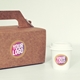 Brown kraft carry box and white takeaway coffee cup, both featuring a circular custom logo sticker with bold text in pink, white, and yellow. Ideal for personalised branding or packaging solutions.