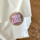 White paper bag sealed with a circular custom logo sticker featuring bold text in pink, white, and yellow on a kraft background. Perfect for personalised branding on takeaway or gift packagi