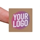 Kraft square label sticker with a custom purple and white logo, shown on a fingertip. The natural brown background of the label gives a rustic and eco-friendly appearance, ideal for personalised packaging