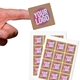 Close-up of a hand holding a small square custom sticker with bold text in pink, white, and yellow on a kraft background. Two sheets of identical stickers are displayed below. Perfect for personalised labelling and branding.