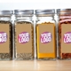 Four glass jars with silver lids, filled with various spices and ingredients, each featuring a personalised kraft label with the text Your Logo in bold pink and white. Ideal for customised kitchen or product branding.