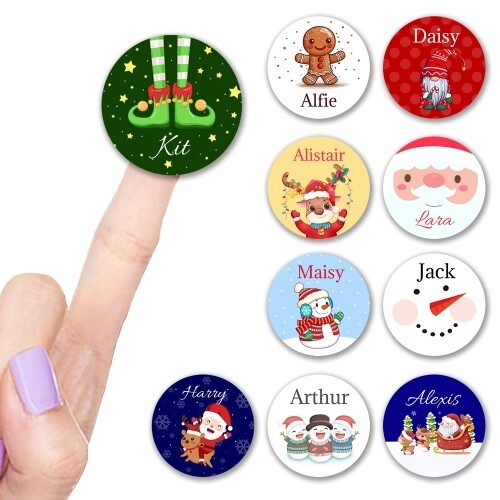 A collection of round personalised Christmas stickers featuring festive designs such as elf legs, Santa, reindeer, gingerbread man, snowman, and gnome. Names include Kit, Alfie, Daisy, Alistair, Lara, Maisy, Jack, Harry, Arthur, and Alexis. A finger with lavender nail polish points to the Kit sticker.