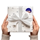 A pair of hands holding a white gift box with silver stars and a white ribbon. The gift features a personalised Christmas sticker with the name Alexis, showing Santa in a sleigh pulled by reindeer on a dark blue background.