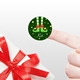 A close-up of a finger holding a round personalised Christmas sticker featuring elf legs in green shoes with red trim. The background is dark green with yellow stars, and the name Kit is written in white. A white gift with a red bow is partially visible.