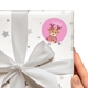 White gift wrapped in star-patterned paper with a white bow, featuring a round personalised Christmas sticker. The sticker shows a cute reindeer named Callie, holding a wrapped present. The sticker has a pink background.