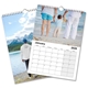 A 2026 wall calendar is shown open to January, featuring a family holding hands on a beach as the top image. The calendar grid below highlights the month of January with large, clear date boxes. Another page beneath shows a scenic image of a man standing by a mountain lake with snowy peaks and pine trees in the background. The calendar is wire-bound with a central hanging hook.