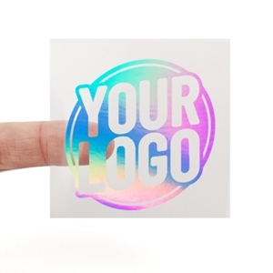 A transparent square sticker with rainbow holographic foil text reading Rainbow Holographic Foiled Stickers. A person's finger is visible behind the sticker, showing its clear background. The foil lettering reflects different colours depending on the angle.