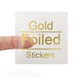 A transparent square sticker with gold foil text reading Gold Foiled Stickers. A person's finger is visible behind the sticker, demonstrating its clear background. The gold foil lettering adds a premium and reflective finish.