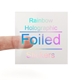 A transparent square sticker with rainbow holographic foil text reading Rainbow Holographic Foiled Stickers. A person's finger is visible behind the sticker, showing its clear background. The foil lettering reflects different colours depending on the angle.