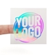 A transparent square sticker with rainbow holographic foil text reading Rainbow Holographic Foiled Stickers. A person's finger is visible behind the sticker, showing its clear background. The foil lettering reflects different colours depending on the angle.