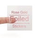 A transparent square sticker with rose gold foil text reading Rose Gold Foiled Stickers. A person's finger is visible behind the sticker, showing its clear background. The foil lettering has a metallic sheen, creating an elegant and premium look.