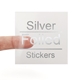 A transparent square sticker with silver foil text reading Silver Foiled Stickers. A person's finger is visible behind the sticker, showing its clear background. The silver foil lettering has a reflective metallic finish for a premium look.