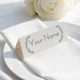Personalised wedding place card with your name in elegant design on folded white cardstock. Positioned on a neatly folded white napkin atop a white plate, with a soft-focus white rose in the background.