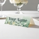 Personalised wedding place card with your name in elegant design on folded white cardstock. Displayed on a white tablecloth with wine glasses, plates, and a floral centrepiece in the softly blurred background.